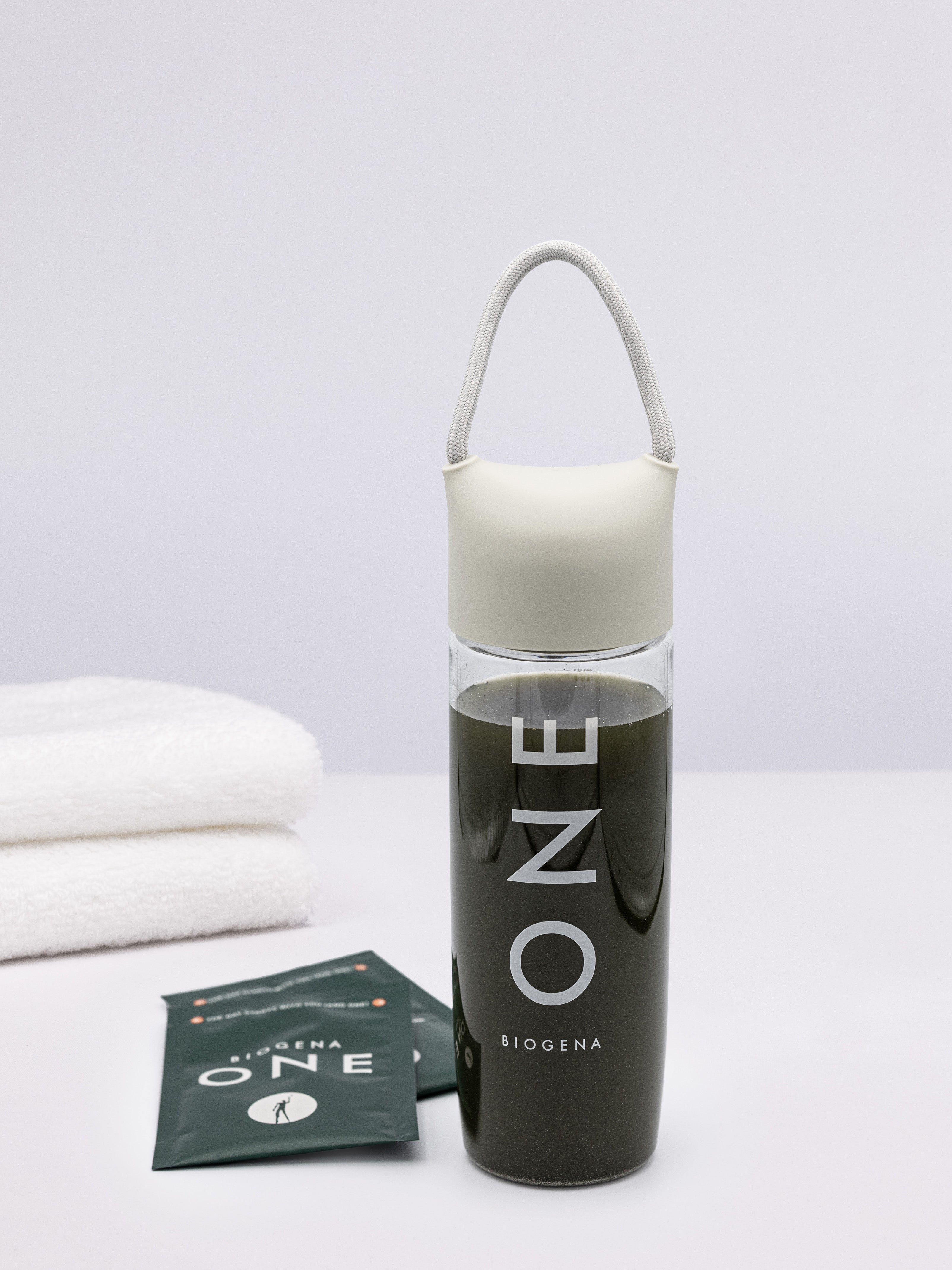 Das BIOGENA ONE All-in-One Supplement-Drink mit Portionsbeutel  und Mixflasche auf einem grauen Untergrund mit weißen Handtüchern seitlich arrangiert.