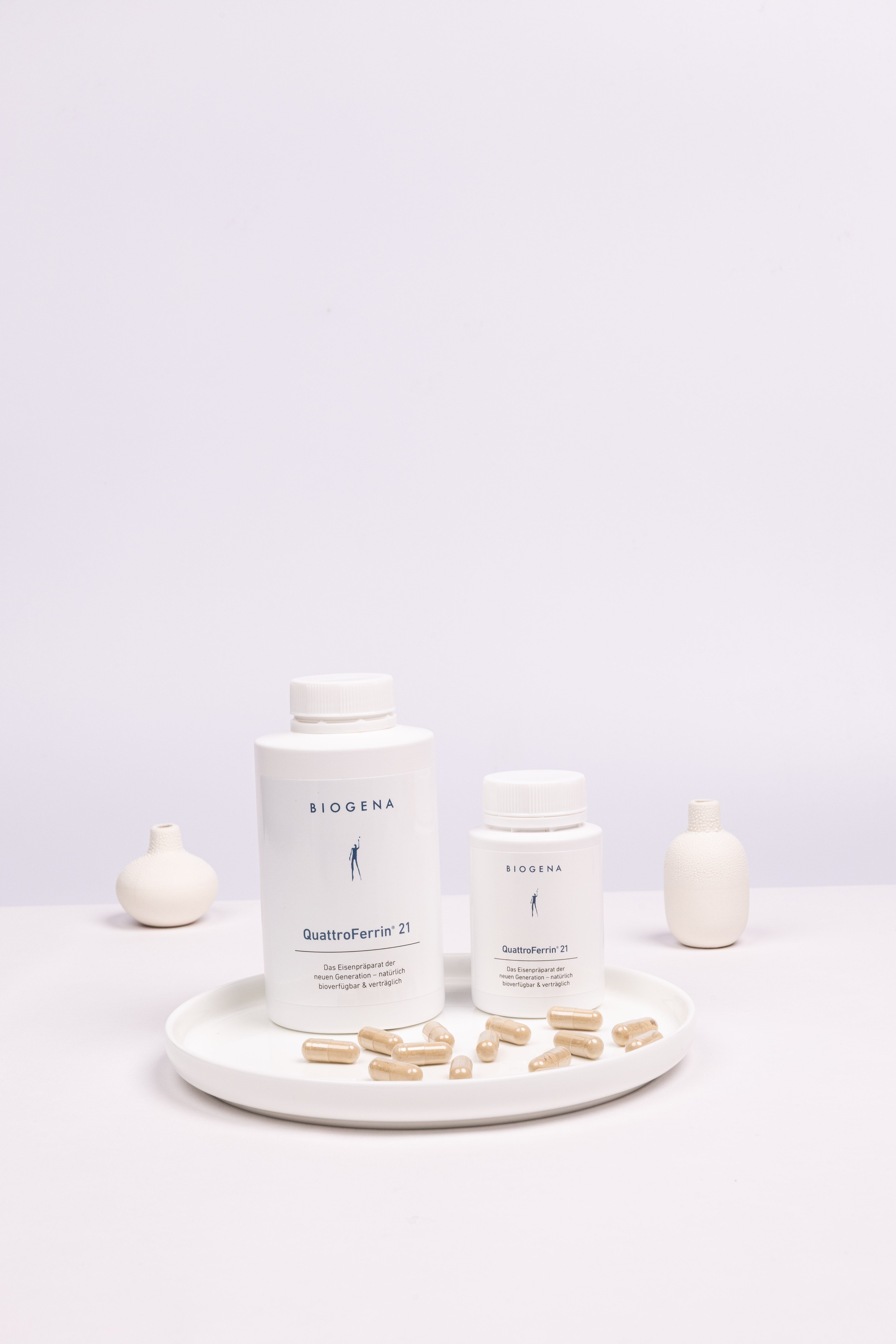 Zwei Dosen QuattroFerrin 21 unterschiedlicher Verpackungsgrößen, stehen auf einem weißen Tablett umgeben von Kapseln.