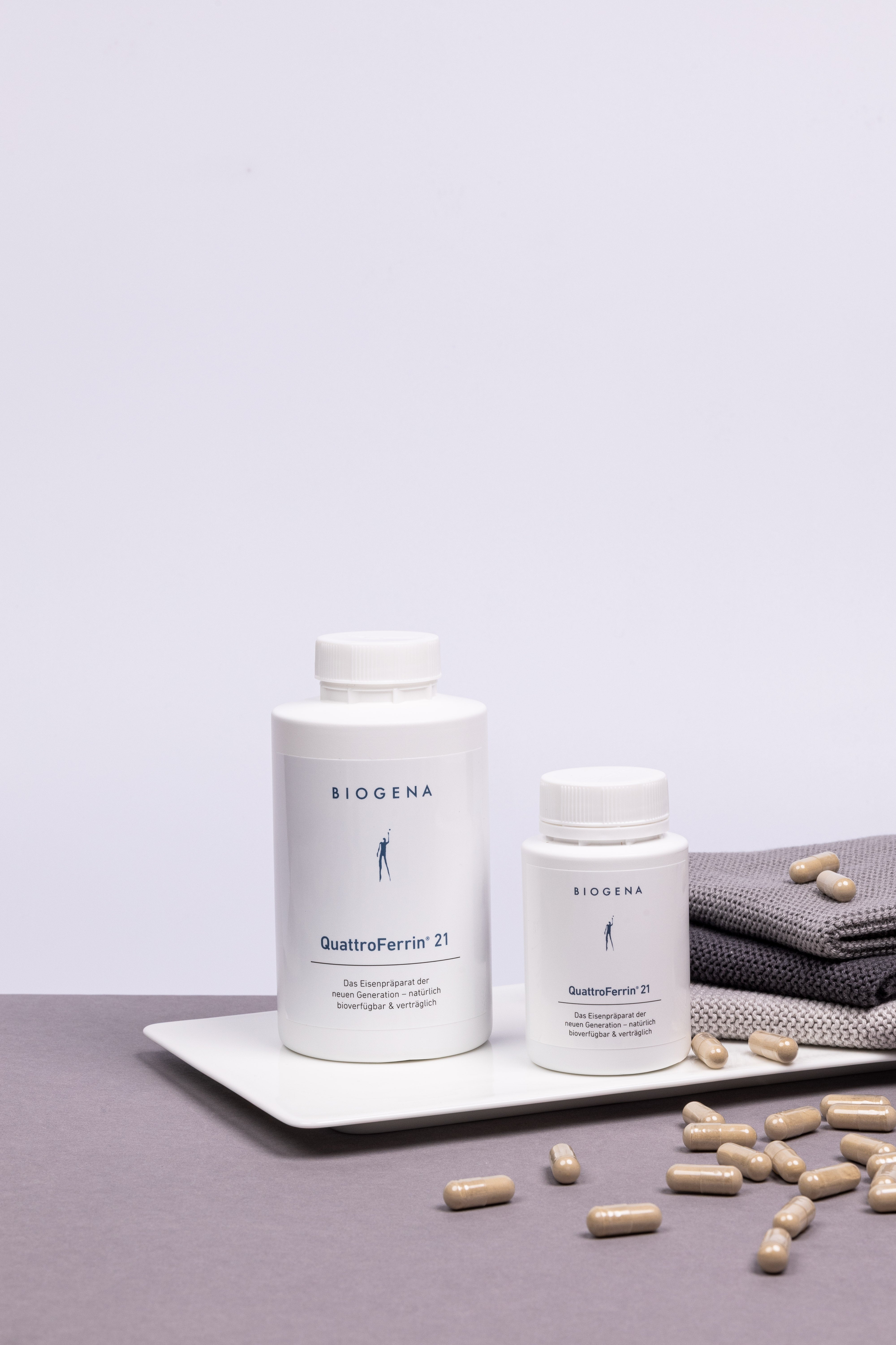 Zwei Dosen QuattroFerrin 21 stehen auf einem Tablett umgeben von Kapseln, rechts daneben ist ein Stapel grauer Handtücher erkennbar.