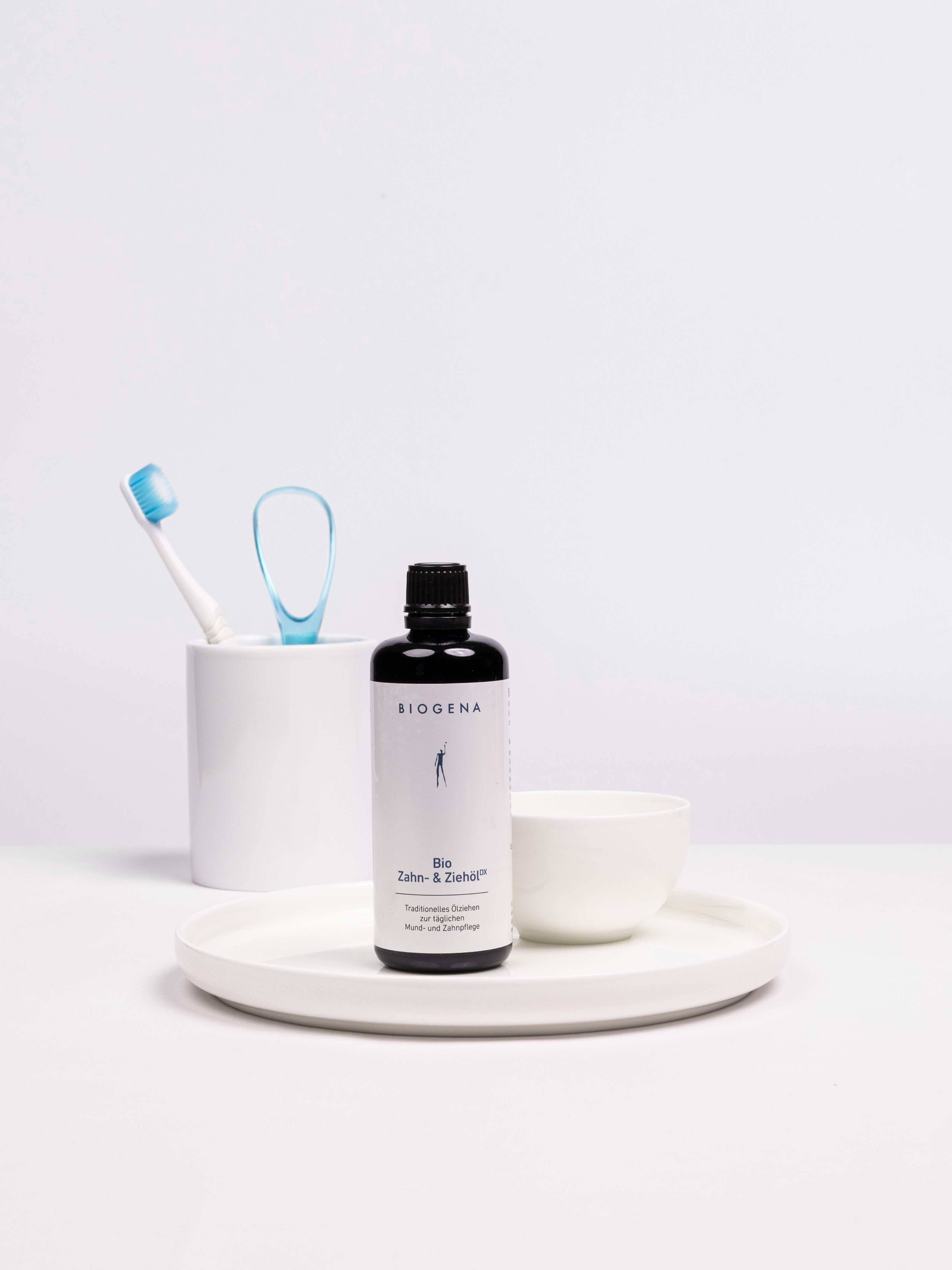 Flasche BIOGENA Zahn- und Ziehöl  auf einem weißen Tablett stehend, links im Hintergrund ist ein Zahnputzbecher mit Zahnbürste und Zungenreiniger erkennbar.