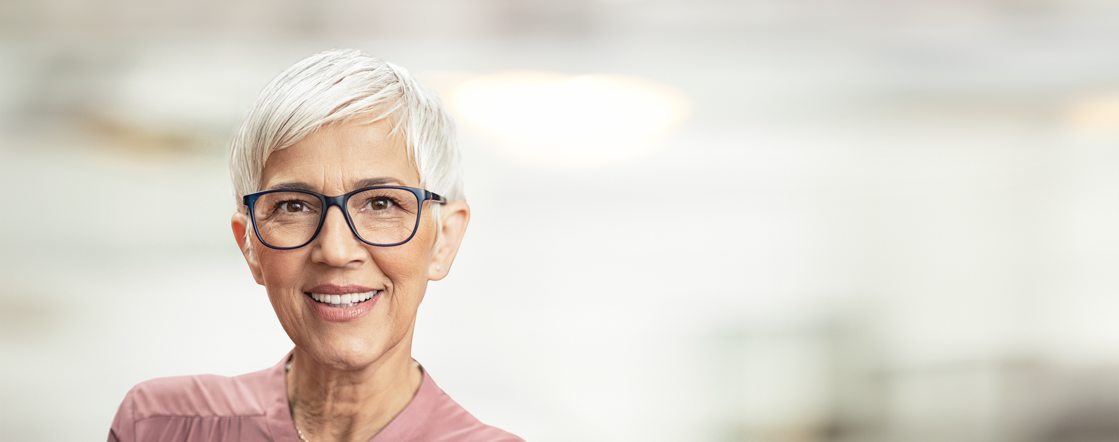 Ältere Frau mit weißen, kurzen Haaren und einer markanten schwarzen Brille, schaut zufrieden und lächelt.