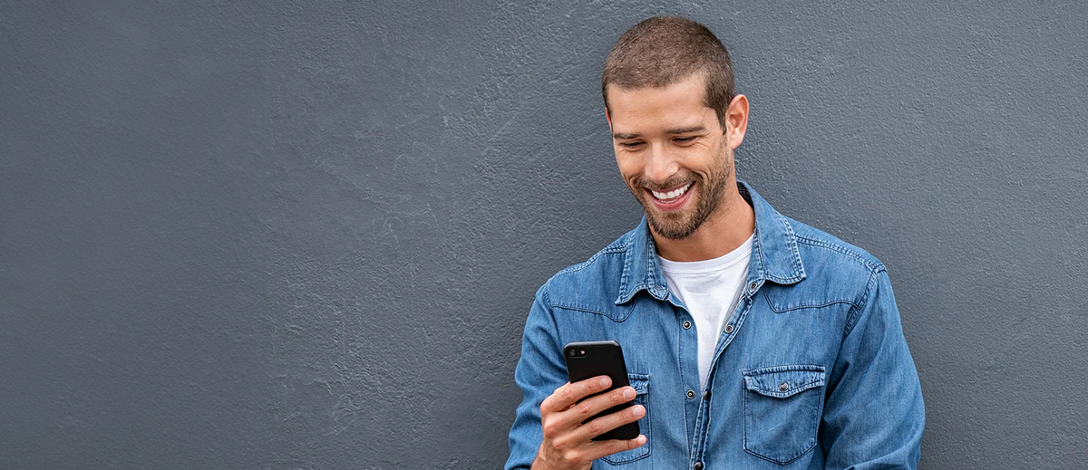 Junger Mann im blauen Jeanshemd, der lächelnd in sein Smartphone schaut.