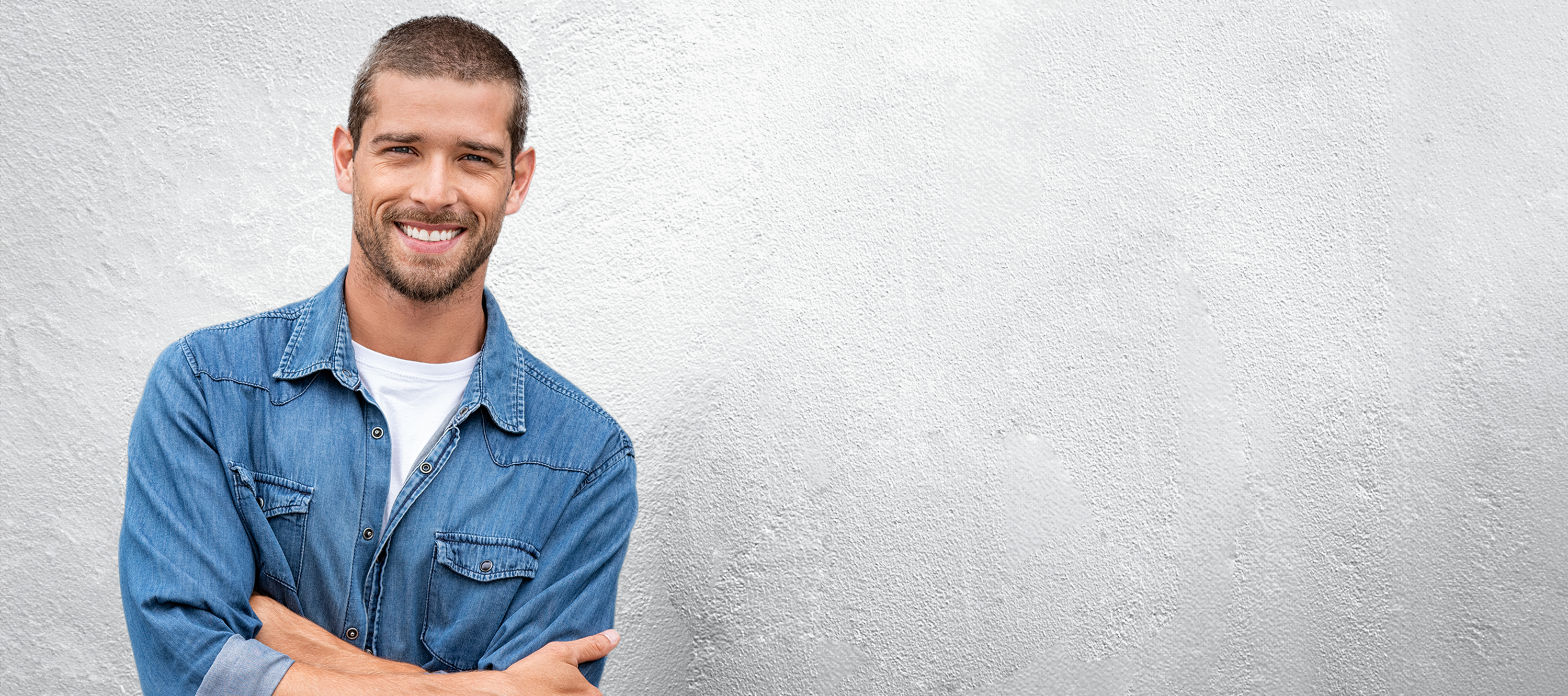 Junger Mann mit kurzen braunen Haaren, im blauen Jeanshemd, lehnt an einer grauen Hauswand. Er hat dabei die Arme verschränkt und lächelt.