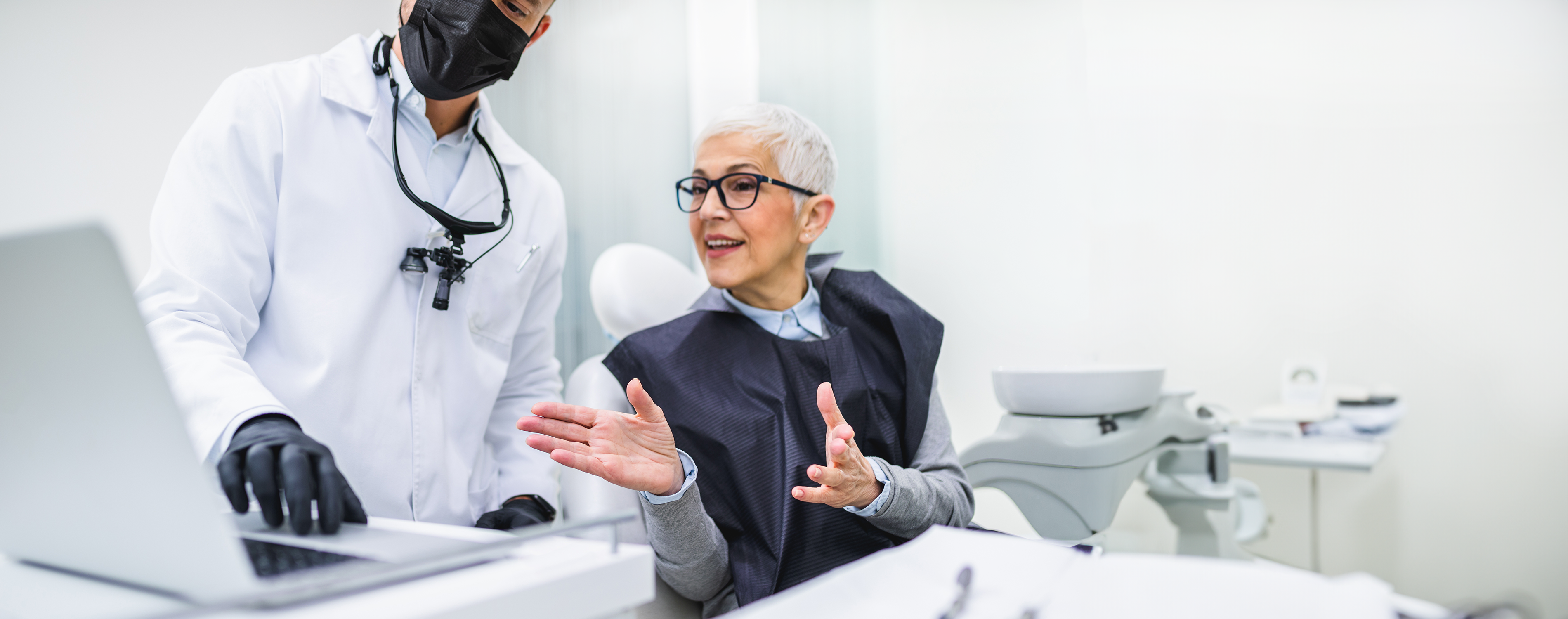 Ältere Patientin sitzt auf dem Behandlungsstuhl einer Zahnarztpraxis und bespricht sich mit ihrem behandelnden Zahnarzt.